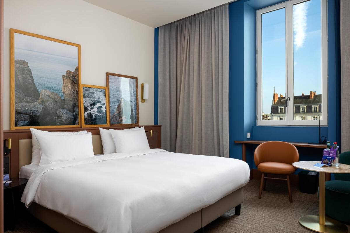 Habitación de hotel con cama, dos cuadros enmarcados, una ventana con cortinas, una silla y una mesita.