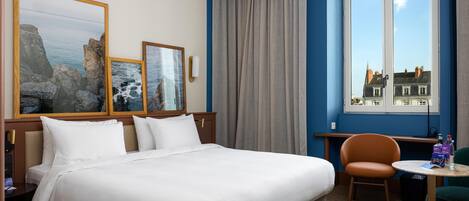 A hotel room with a bed, two framed pictures, a window with curtains, a chair, and a small table.