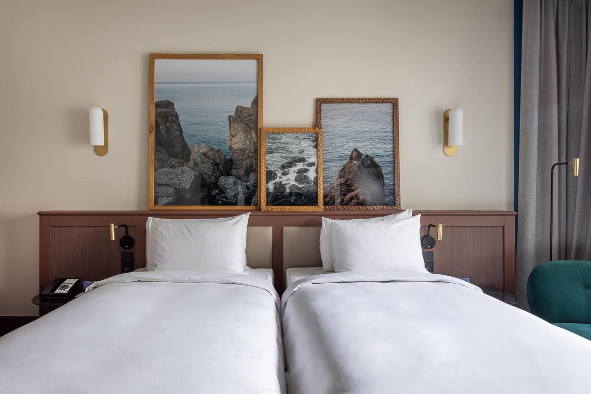 Habitación de hotel con dos camas, cabecero de madera, teléfono y una obra de arte que representa un paisaje costero.
