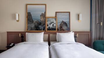 A hotel room with two beds, a wooden headboard, a telephone, and artwork depicting coastal scenery.