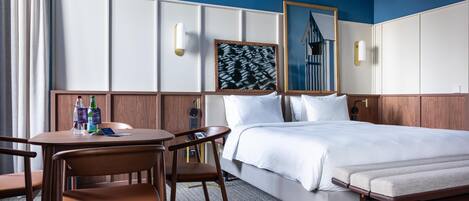 Une chambre d’hôtel comprenant un lit, un bureau, deux chaises, un banc, un miroir et une œuvre d’art encadrée accrochée au mur.