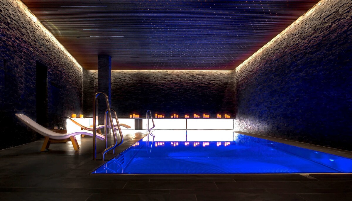 Dark room with a glowing blue pool and lounge chairs. Candles line the room