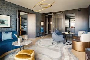 A modern living room with a blue sofa, a gold ottoman, a round coffee table, and a grey armchair. There is a chandelier, a lamp, and a framed picture on the wall.
