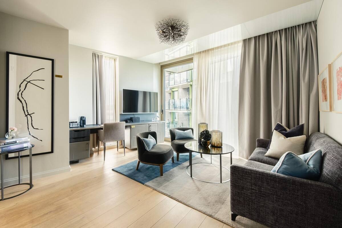 Uma sala de estar moderna com um sofá, duas poltronas, uma mesa de centro e uma televisão. Há uma área de jantar com mesa e cadeiras, e uma cozinha ao fundo.