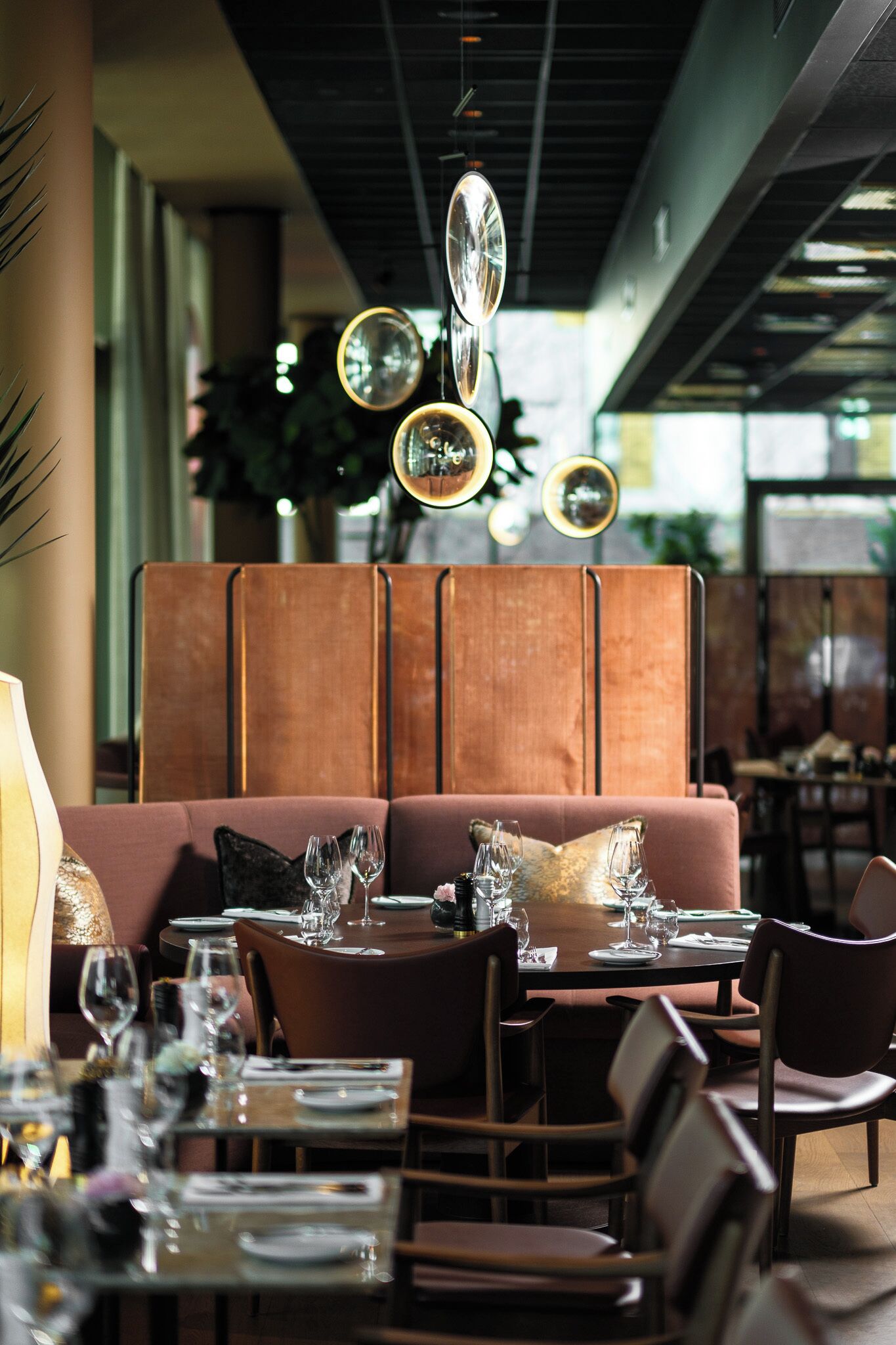 Interior de restaurante com design moderno, apresentando uma longa mesa de jantar posta com copos e talheres, área de estar com assentos acolchoados e iluminação pendente.