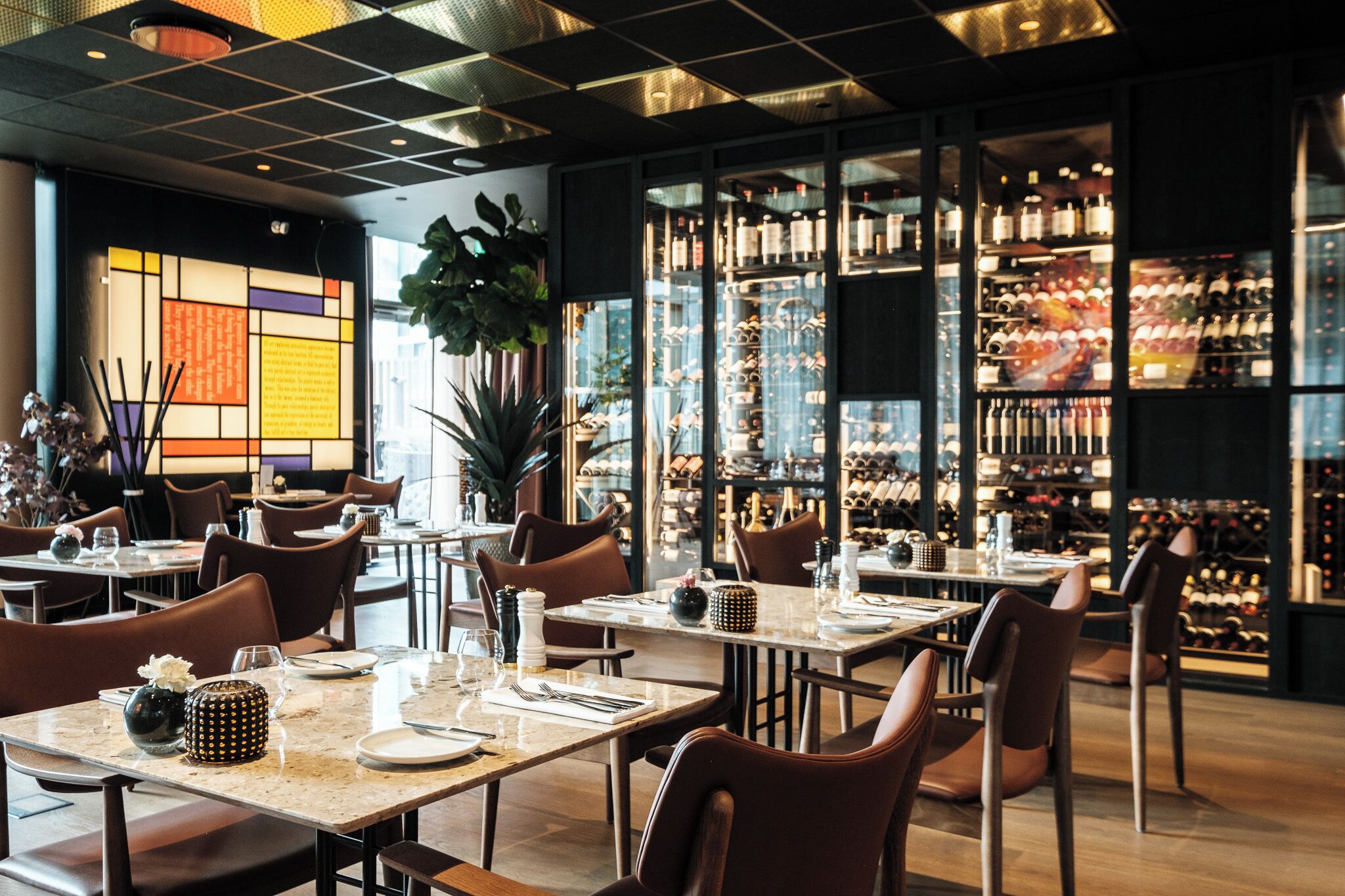 A restaurant interior with tables set for dining, a wine rack in the background, and a large plant in the center.