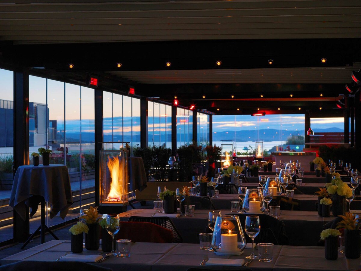 Dining room set with candles and flames, lined with floor to ceiling windows with views of the Oslo harbor at sunset