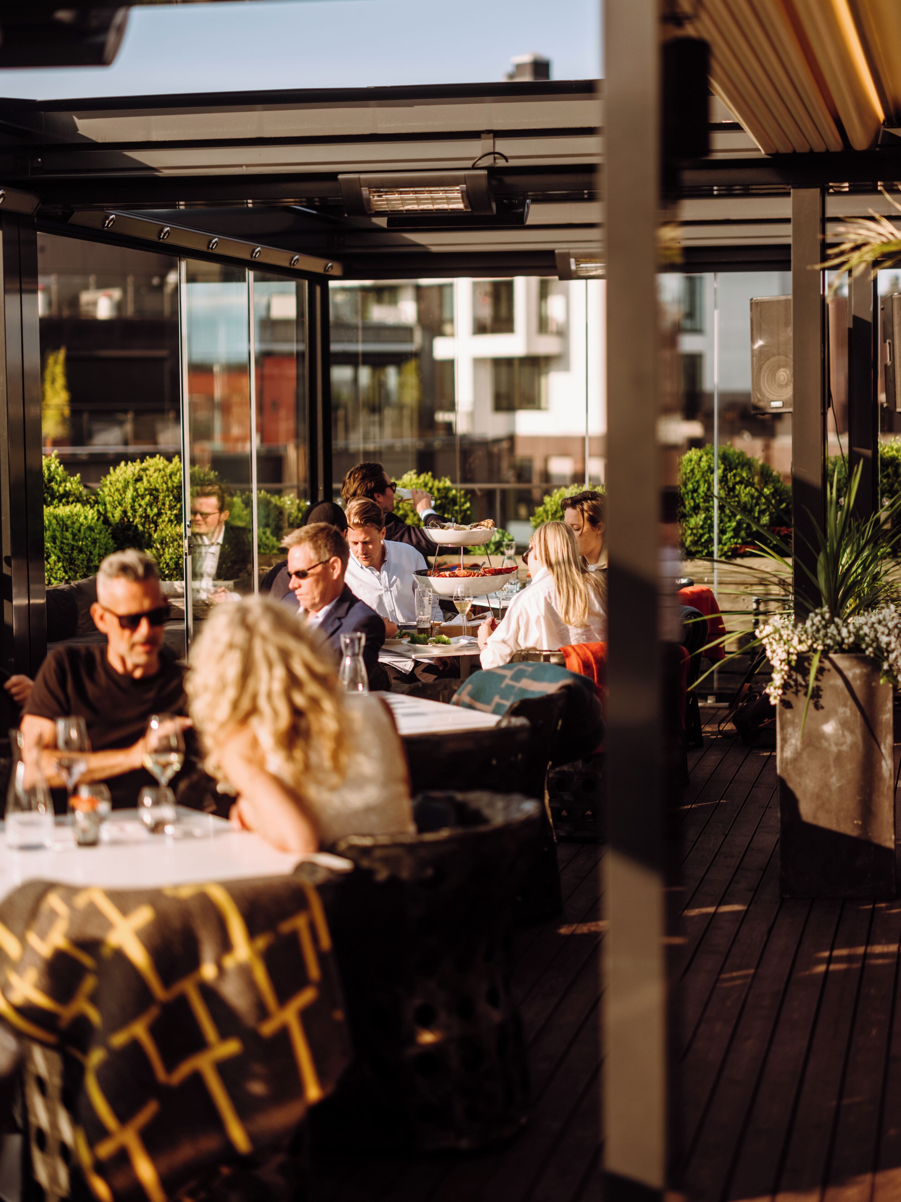 Een terras met tafels en stoelen, grote ramen en potplanten.