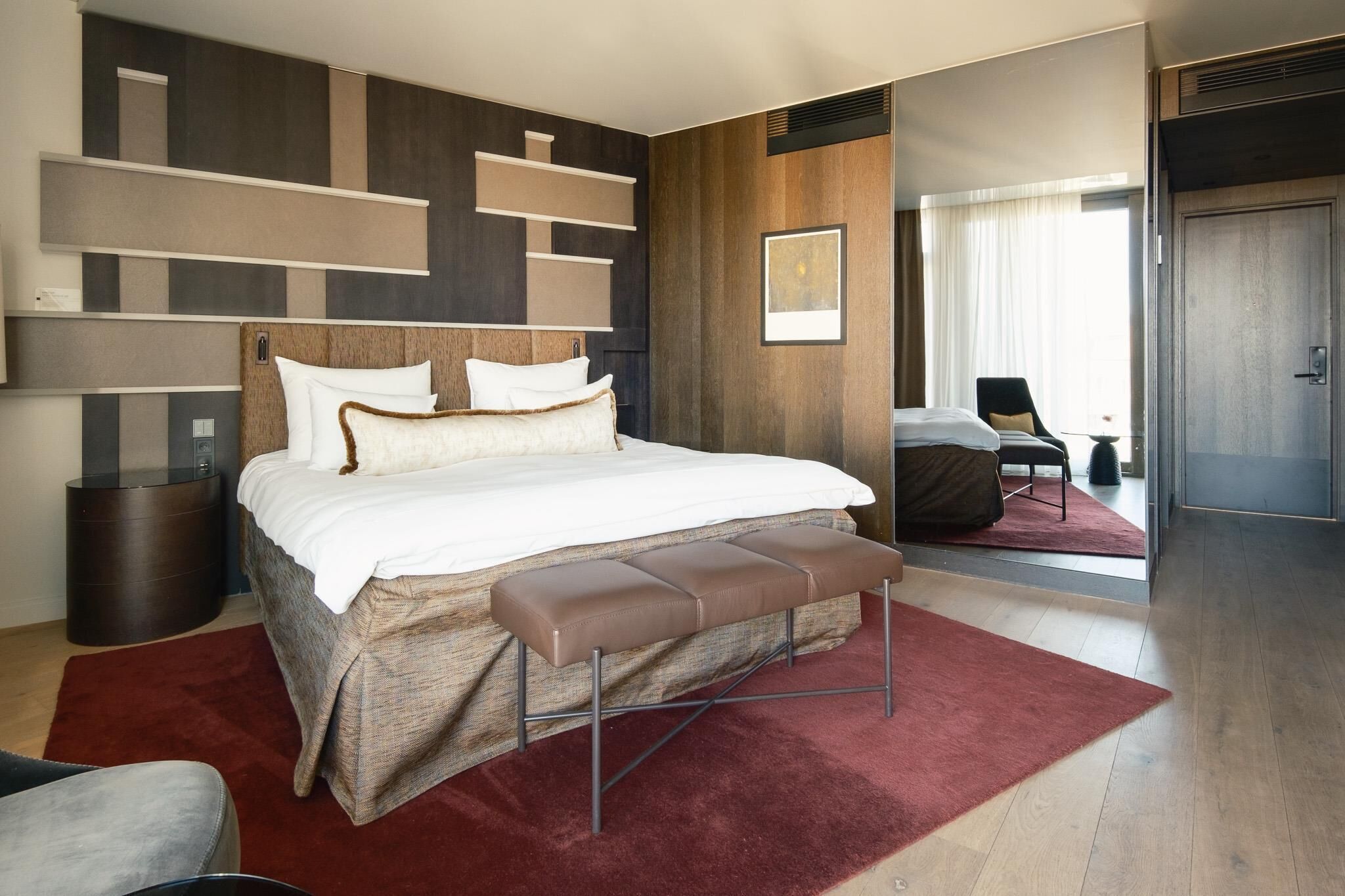 A modern hotel room with a large bed, a bench, a chair, and a red rug.