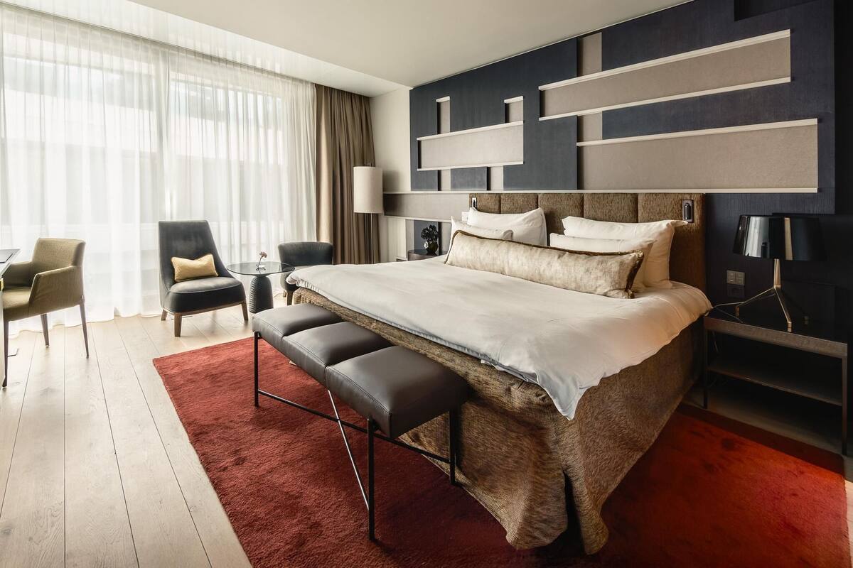 Quarto de hotel moderno com uma cama grande, um tapete vermelho, área de estar com cadeiras e uma janela ampla com cortinas.