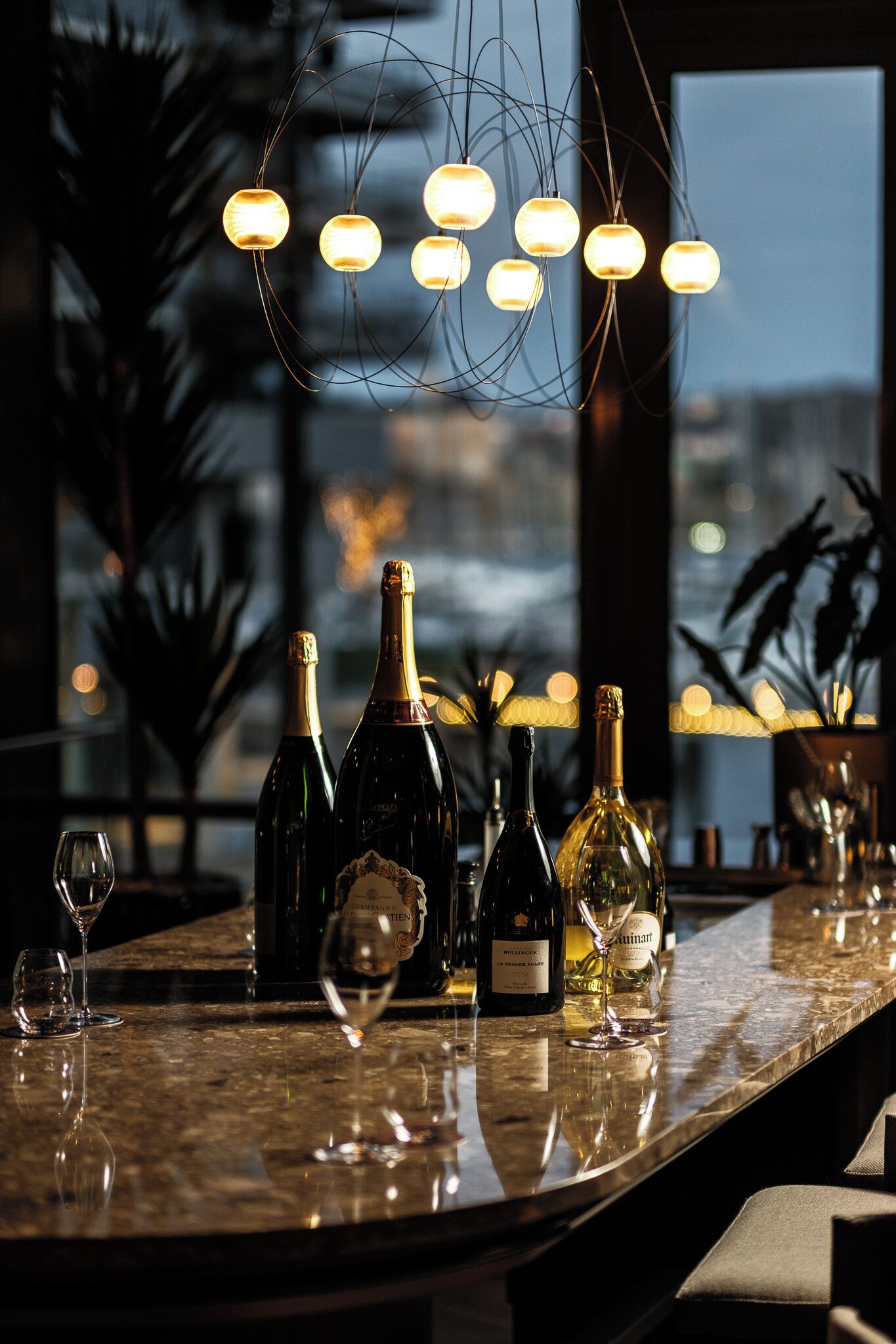 A bar area with multiple bottles of wine and glasses, a modern light fixture, and a view of the cityscape through large windows.