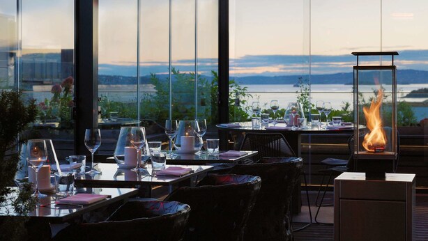 An outdoor dining area with a view of a lake and mountains, equipped with tables, chairs, and a fire feature.
