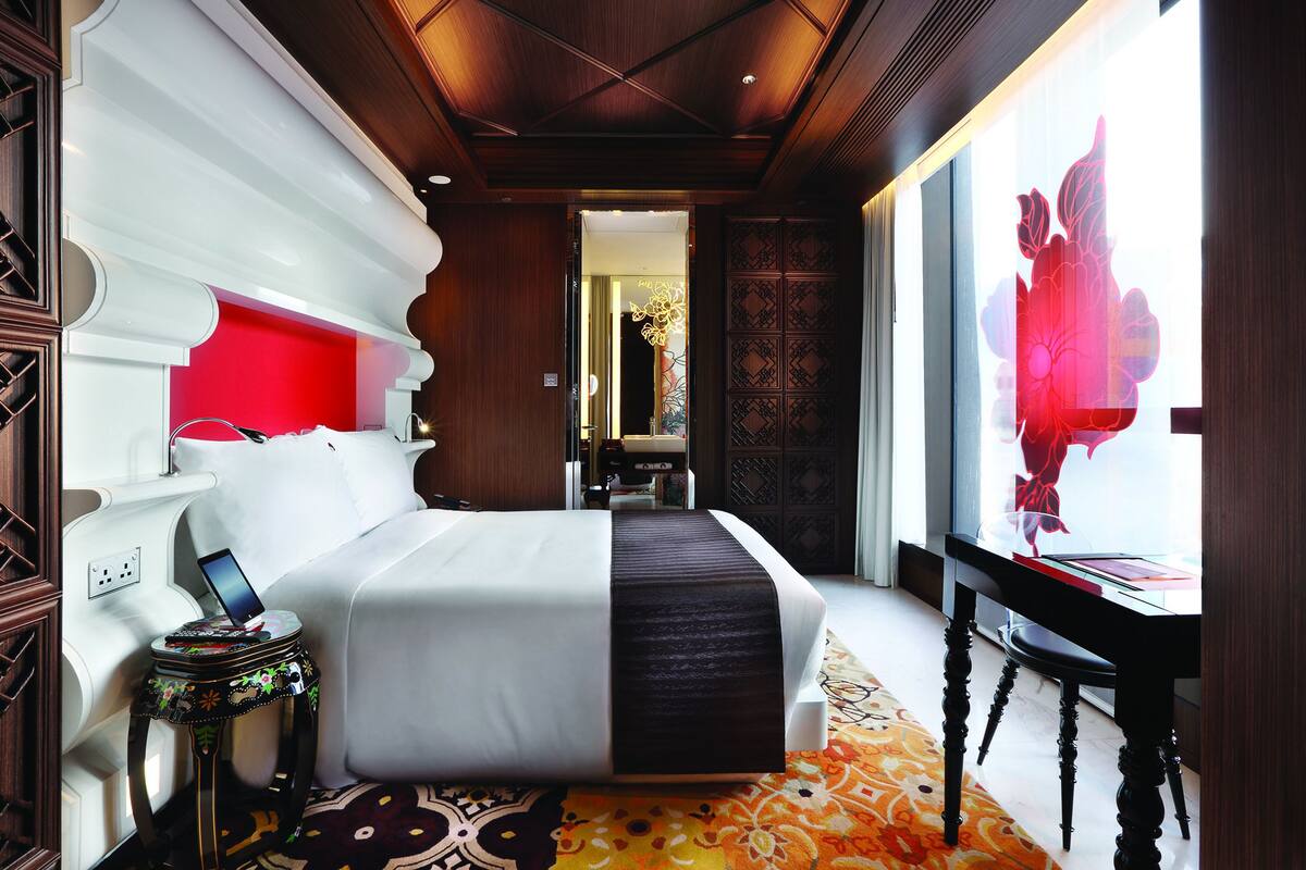 A hotel room with a large bed, a bedside table with a phone, a desk with a chair, and a view of a red floral design.