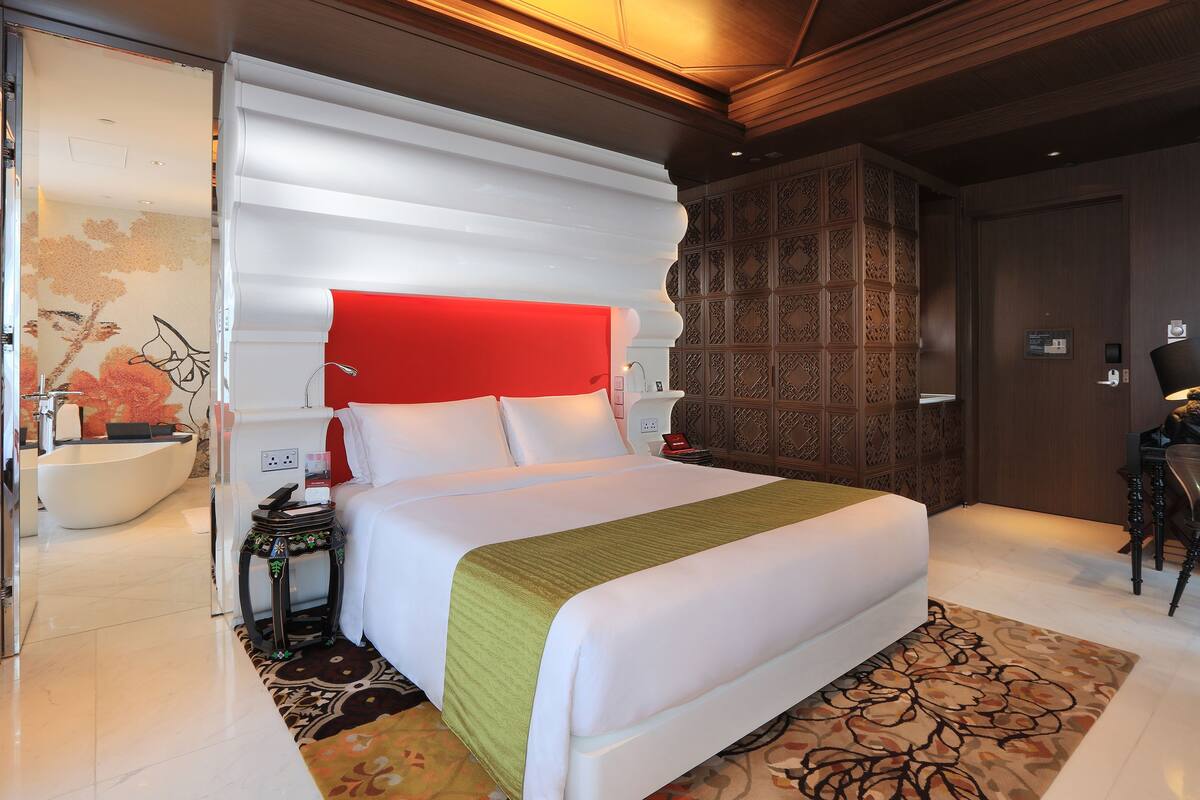 A modern hotel room with a large bed, a red accent wall, a decorative wall panel, a bathroom with a white bathtub, and a patterned rug.