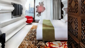 A modern hotel room with a large bed, a red chair, a TV, and a city view.