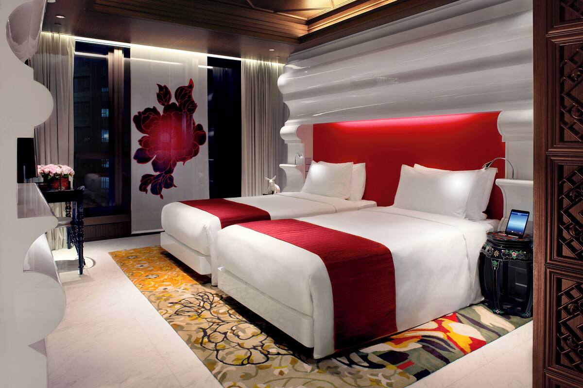 A modern hotel room with two beds, a red accent wall, and a decorative wall piece.