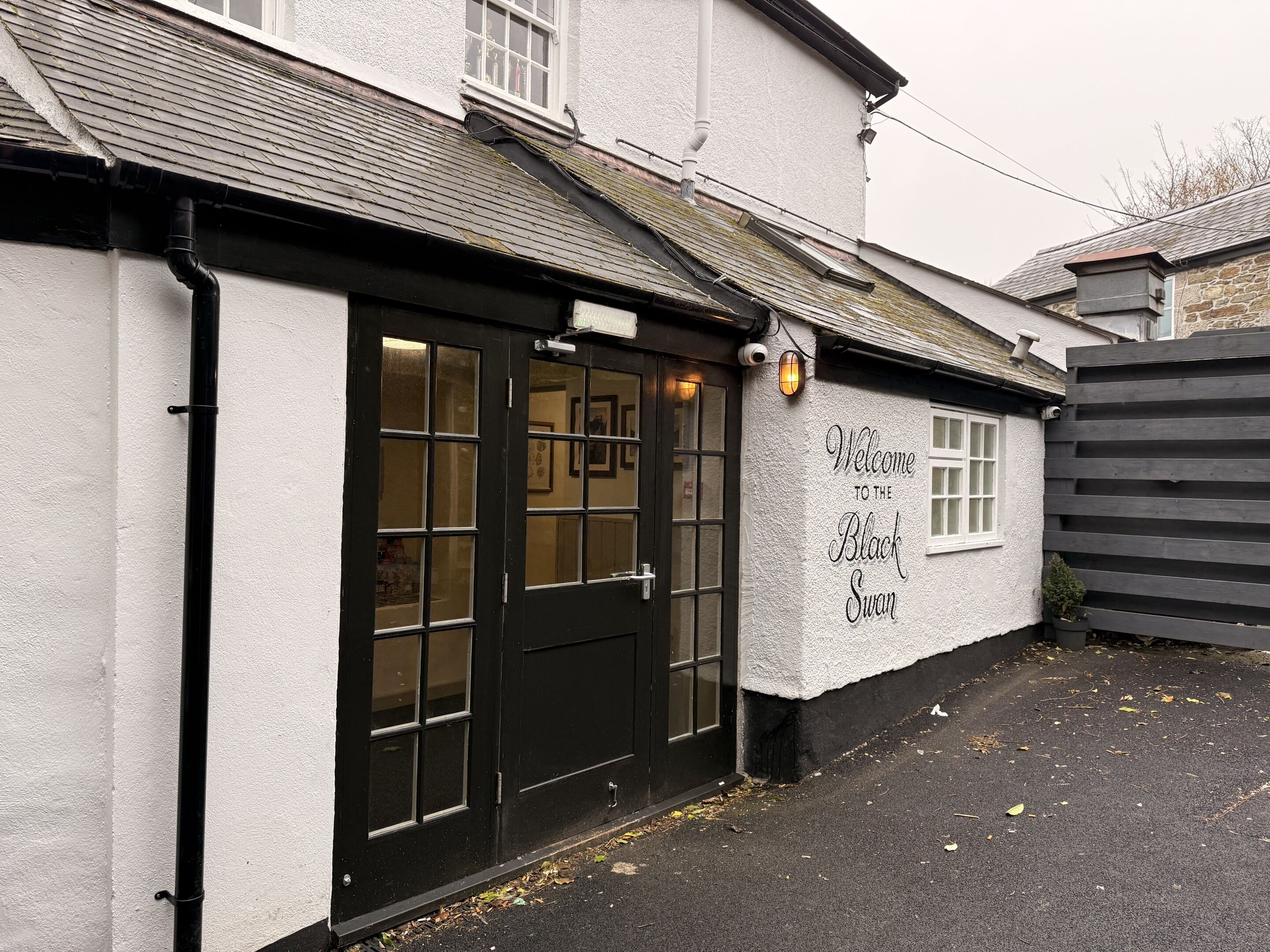 A building with a black door and windows, a sign reading 'Welcome to the Black Swan,' and a small garden area with a fence.