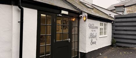 A building with a black door and windows, a sign reading 'Welcome to the Black Swan,' and a small garden area with a fence.