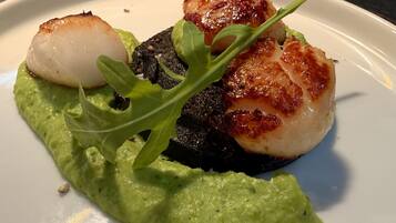 Un plato con vieiras selladas, un trozo de carne oscura y una salsa verde, decorado con rúcula.
