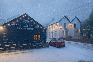 A snow-covered building with a sign for Gweek Village Stores & Farm Shop, outdoor seating, and a parked car. - The Black Swan (Helston)