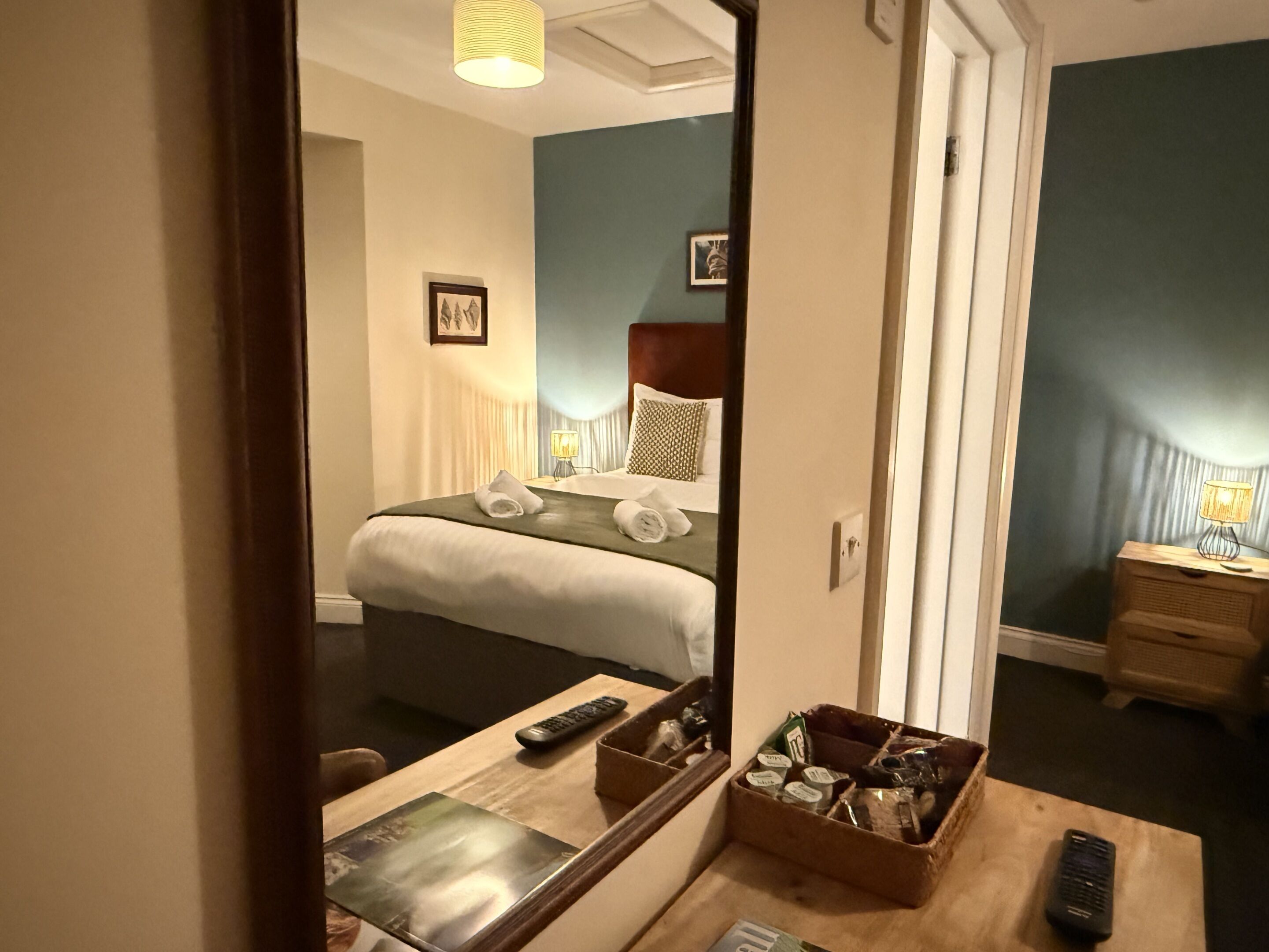A hotel room with a bed, bedside table, lamp, and a mirror reflecting the room.