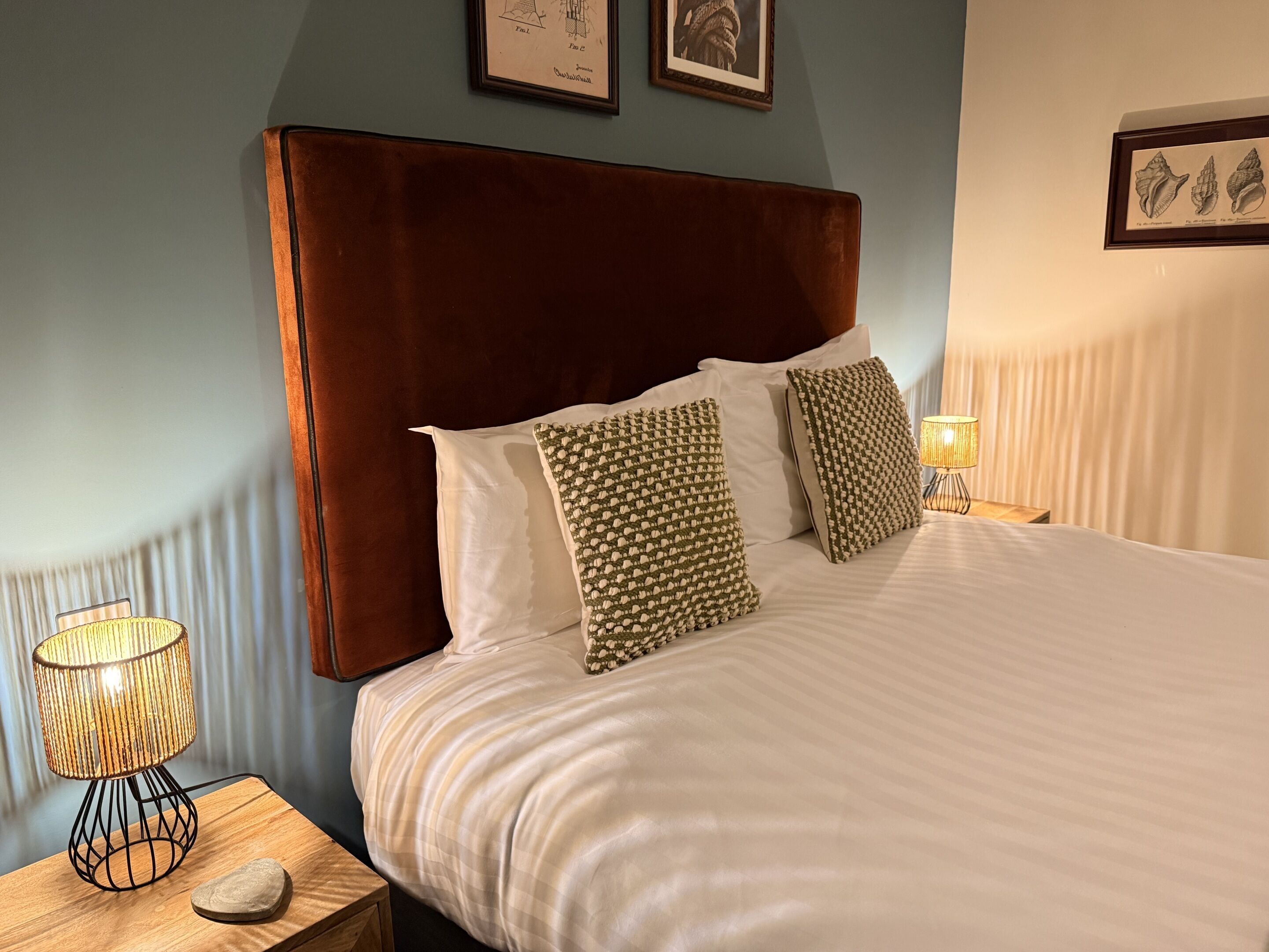 A neatly made bed with a brown headboard, white linens, and patterned pillows. There is a bedside lamp and a framed picture on the wall.