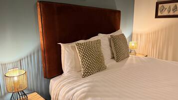 A neatly made bed with a brown headboard, white linens, and patterned pillows. There is a bedside lamp and a framed picture on the wall.