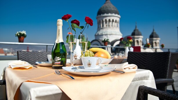 A table set with a bottle of Cinzano, two glasses, a cup, and a bowl of fruit, with a view of a domed building in the background. - Hotel Beyfin (Cluj-Napoca)