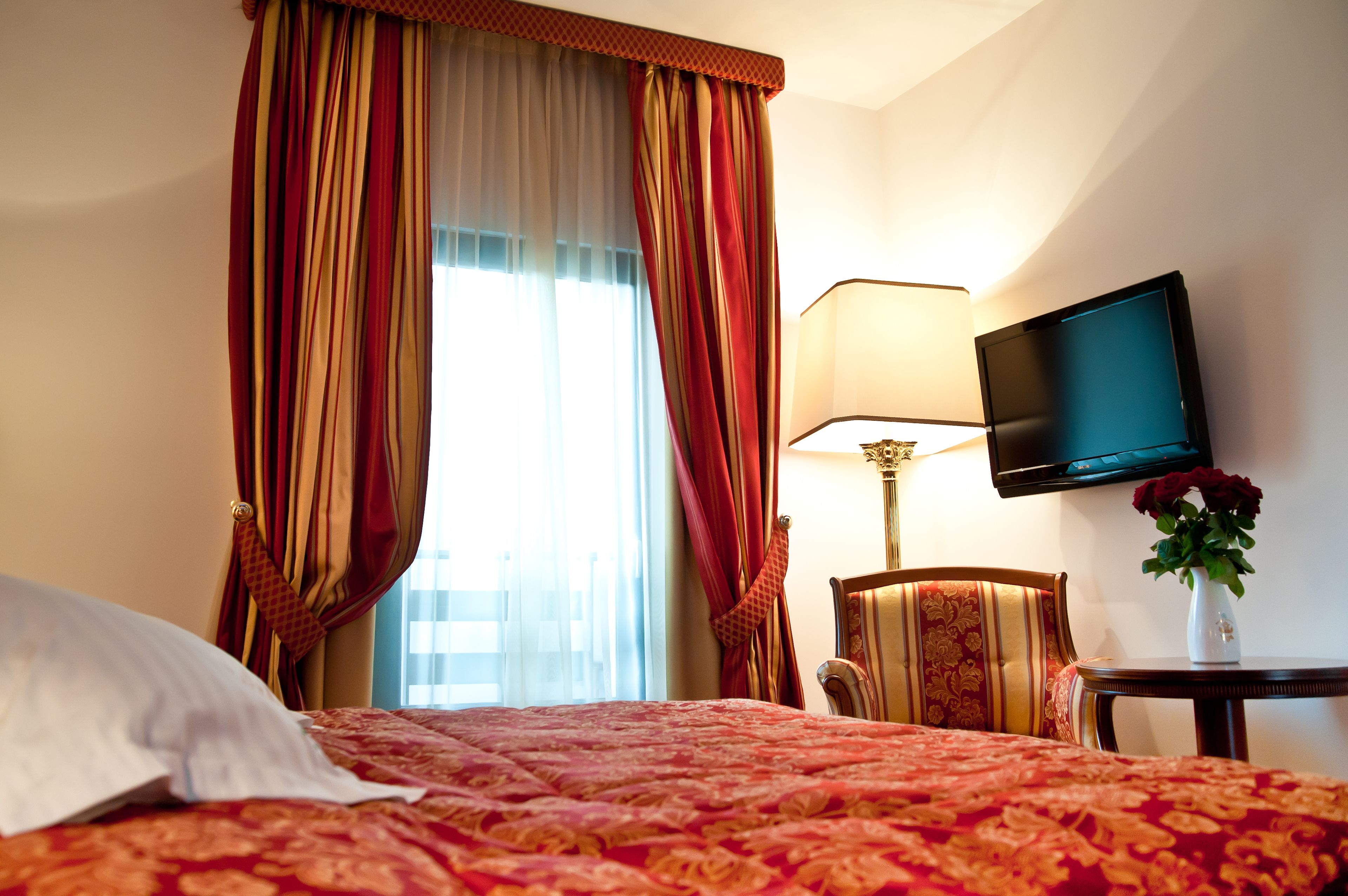 A hotel room with a bed, curtains, a television, a lamp, a chair, and a vase with flowers.