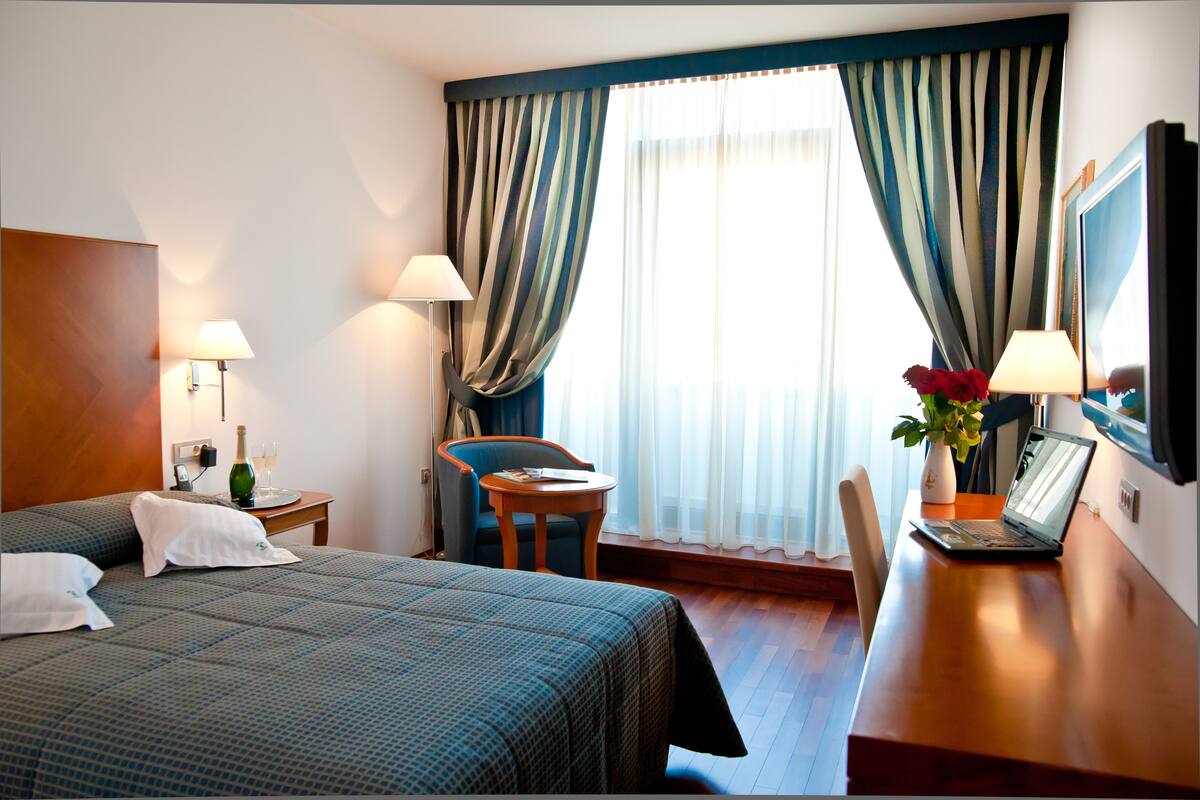 A hotel room with a bed, a desk, a chair, a lamp, a TV, and a window with curtains.