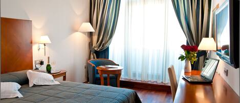 A hotel room with a bed, a desk, a chair, a lamp, a TV, and a window with curtains.