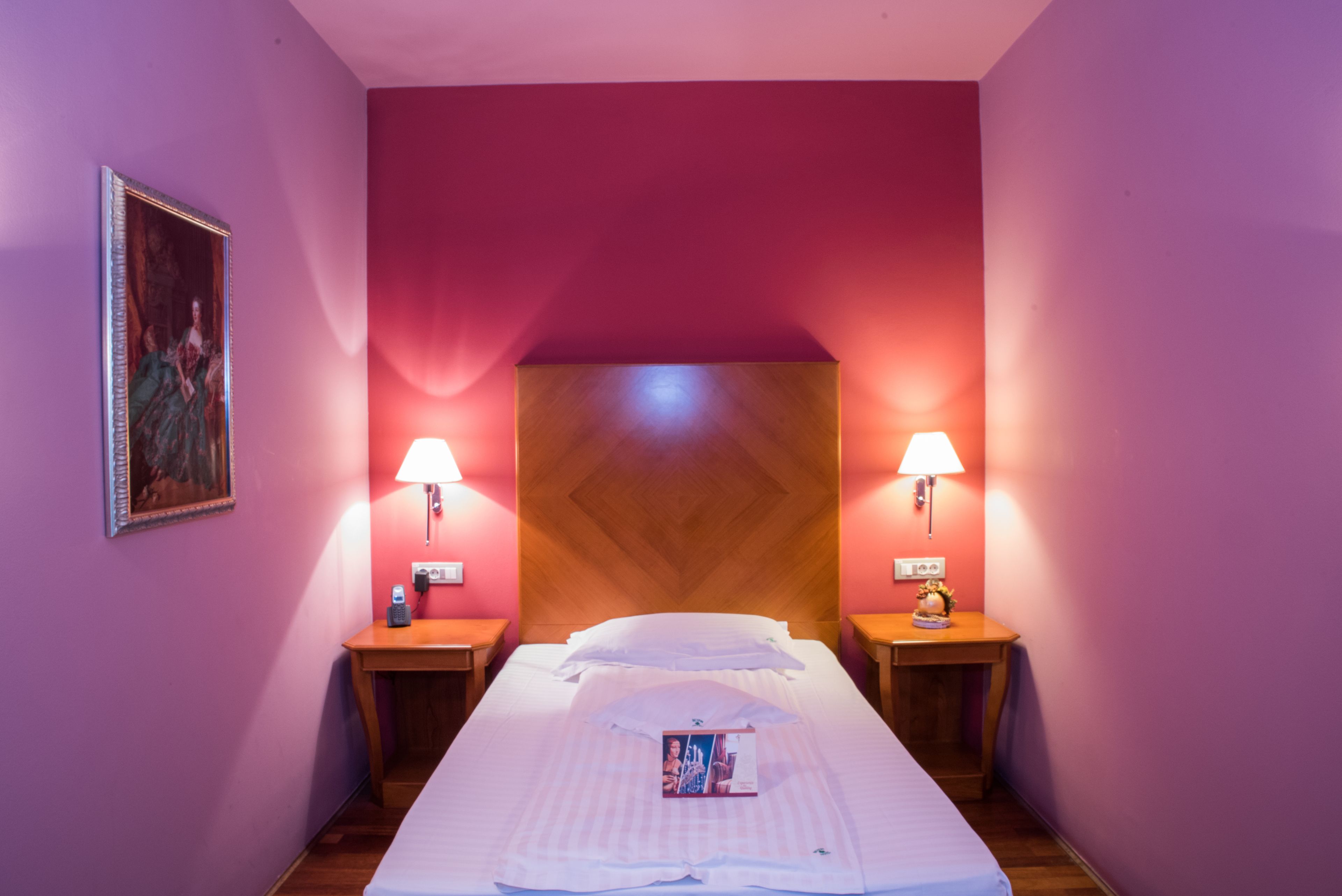 Habitació d'hotel amb un llit, dues taules de nit, una estantalla i una làmpada penjada a la paret, a més d'una fotografia emmarcada.