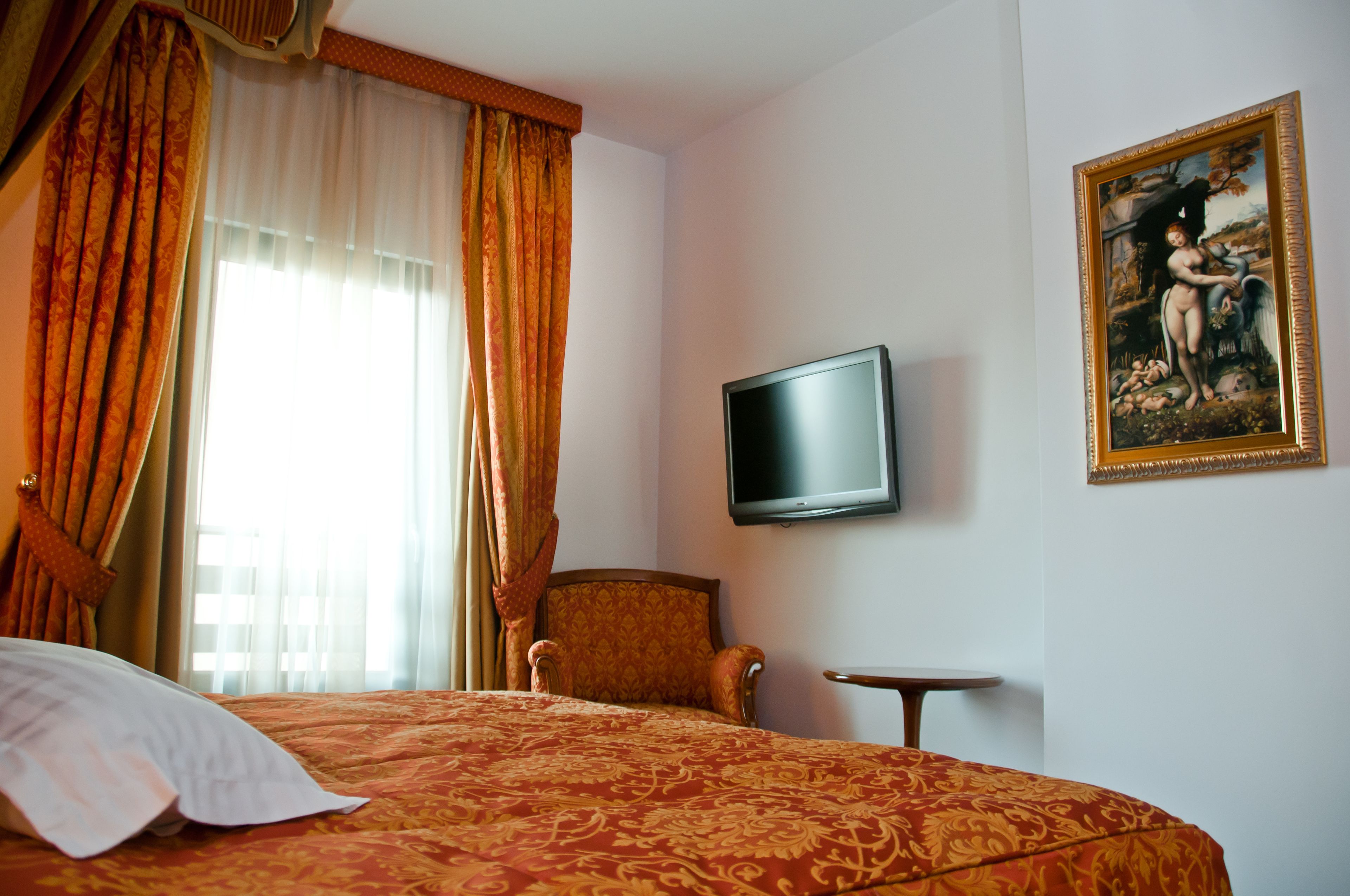A hotel room with a bed, a chair, a small table, a TV, and a painting on the wall.