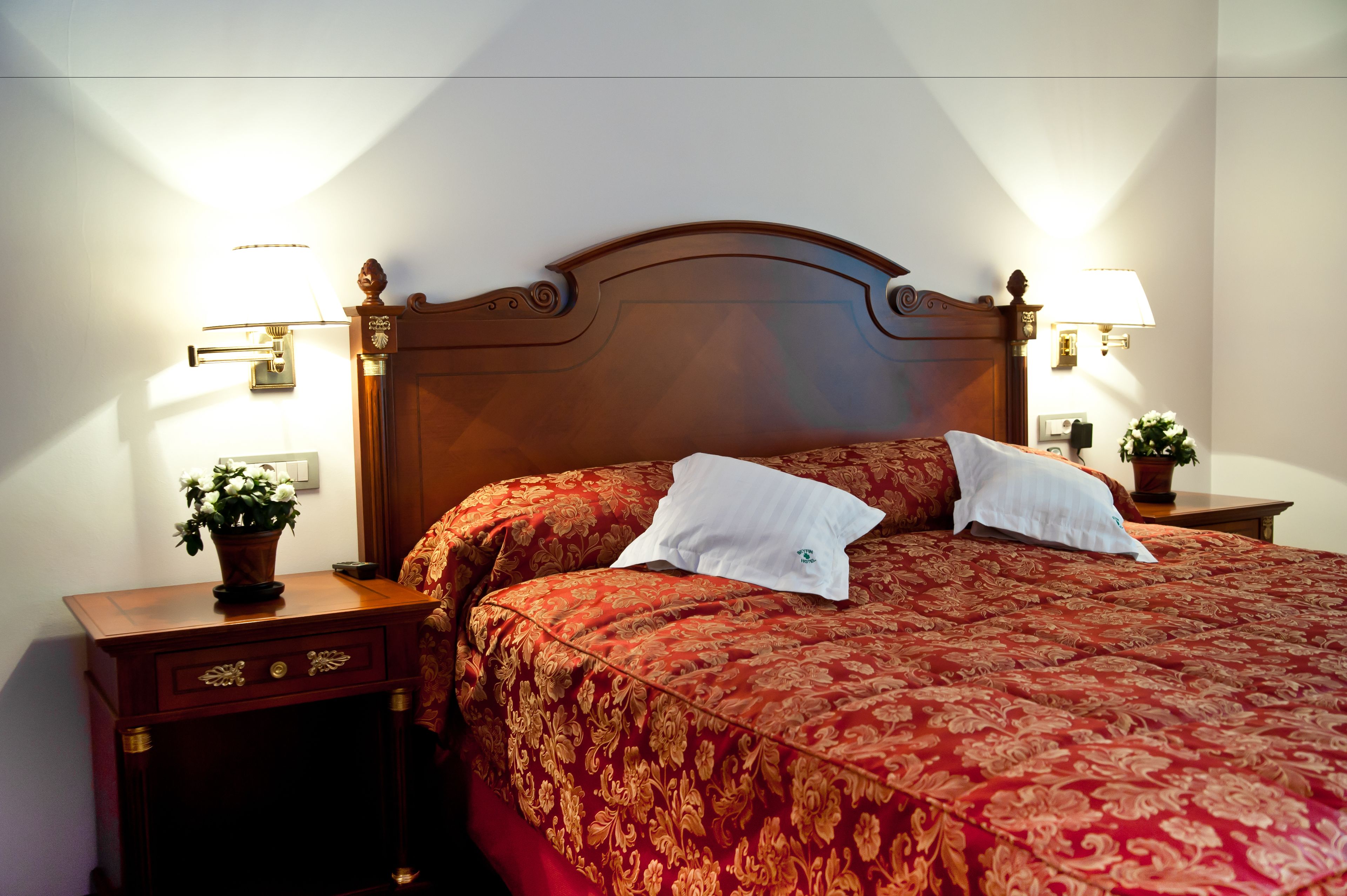 A neatly made bed with a red patterned bedspread and two white pillows, flanked by two bedside tables with lamps and a small potted plant on each.