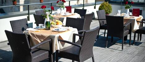 Una terraza en la azotea con mesas dispuestas para comer, con sillas de mimbre y vistas a edificios al fondo.