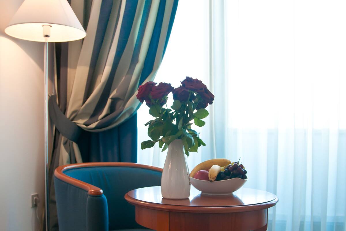 A room with a blue armchair, a wooden side table with a vase of flowers and a bowl of fruit, a floor lamp, and a view through sheer curtains.