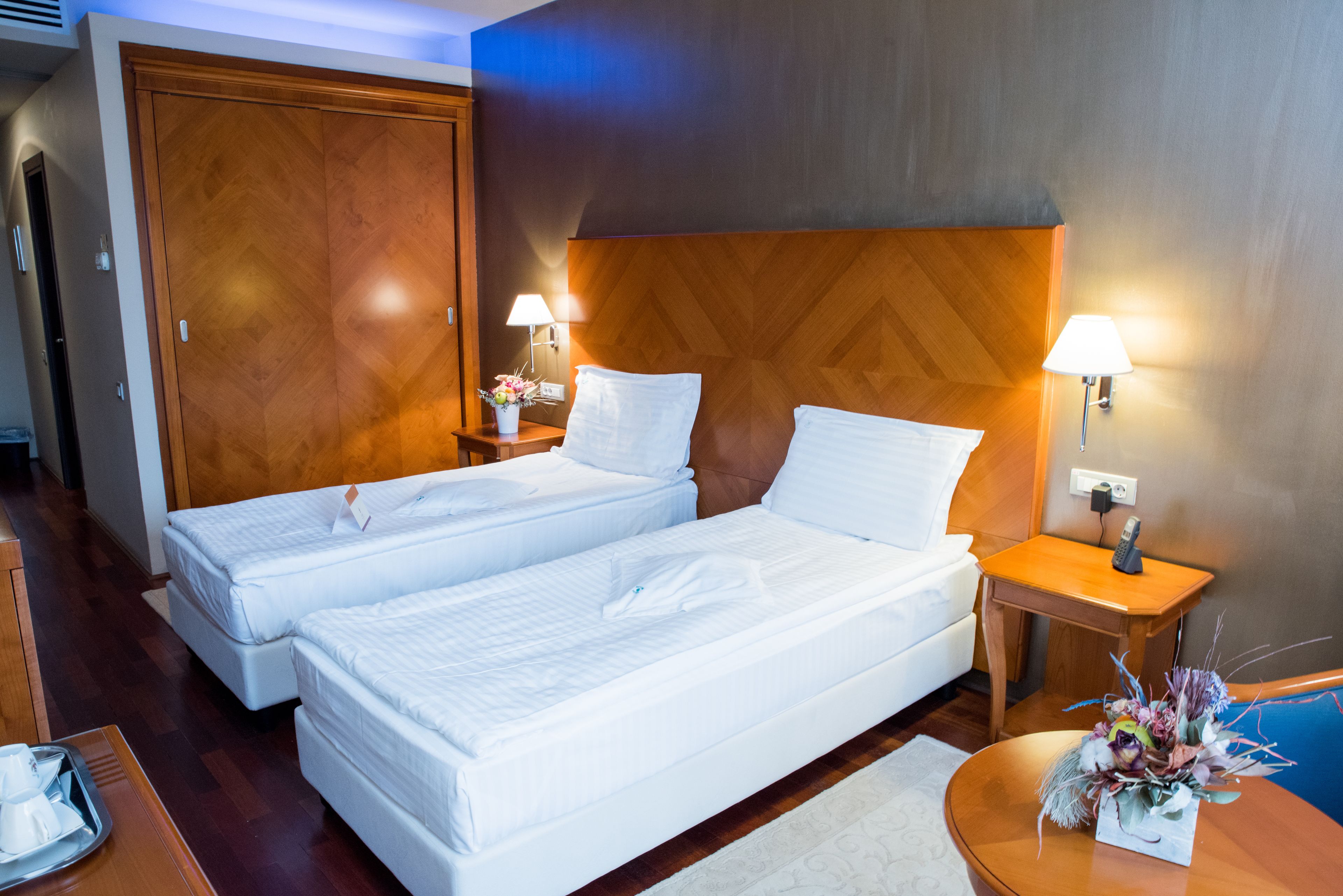 Ein Hotelzimmer mit zwei Betten, einem hölzernen Kopfteil, Nachttischlampen, einem kleinen Tisch mit einer Vase und einem Nachttisch aus Holz.