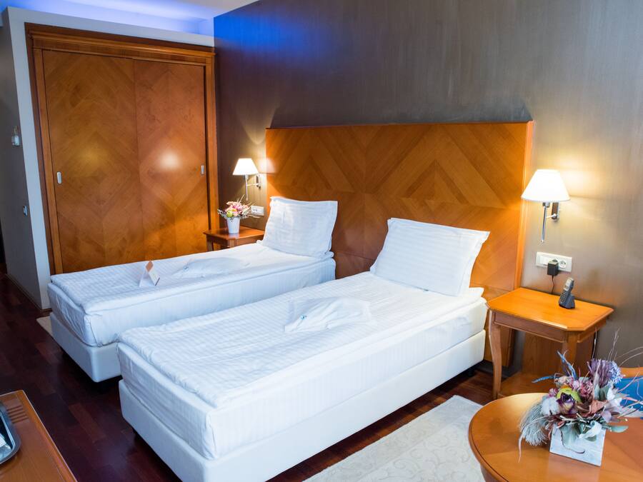 Ein Hotelzimmer mit zwei Betten, einem hölzernen Kopfteil, Nachttischlampen, einem kleinen Tisch mit einer Vase und einem Nachttisch aus Holz.