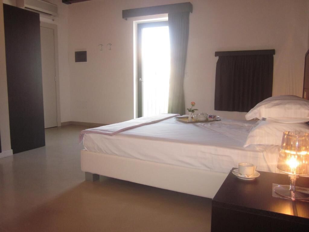 A hotel room with a bed, a bedside table with a cup and a glass, a window with curtains, and a door.