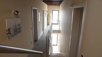A narrow hallway with a staircase leading to a landing. There are doors on both sides, a painting on the left wall, and a light fixture on the ceiling.
