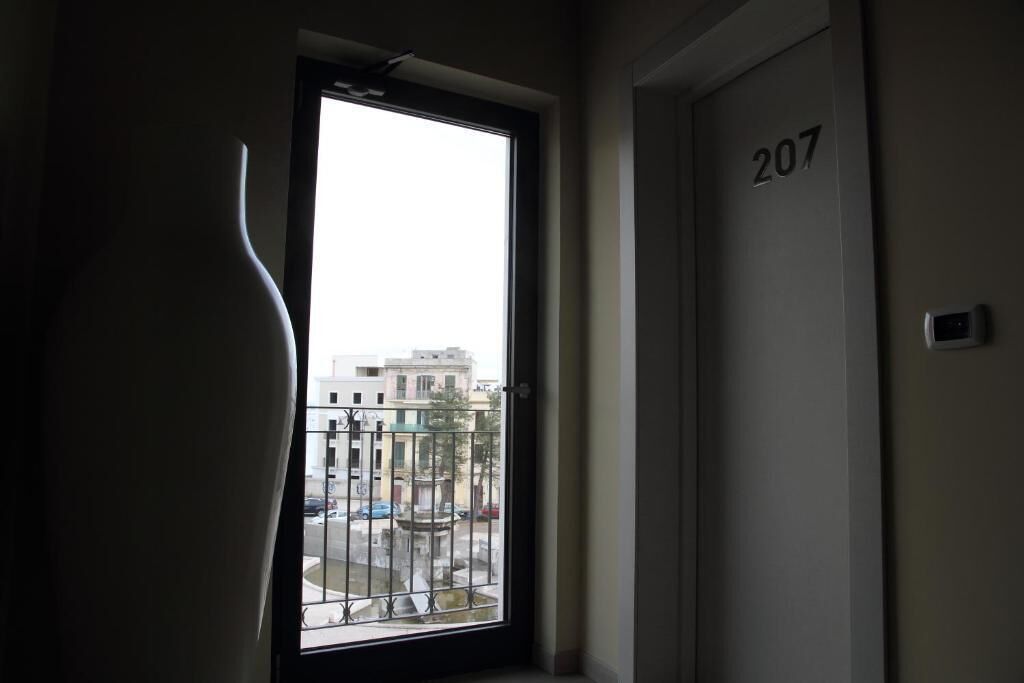 Ein Zimmer mit einer Tür, auf der 207 steht, einem Fenster mit Blick auf Gebäude und eine Straße sowie einer großen Vase auf der linken Seite.