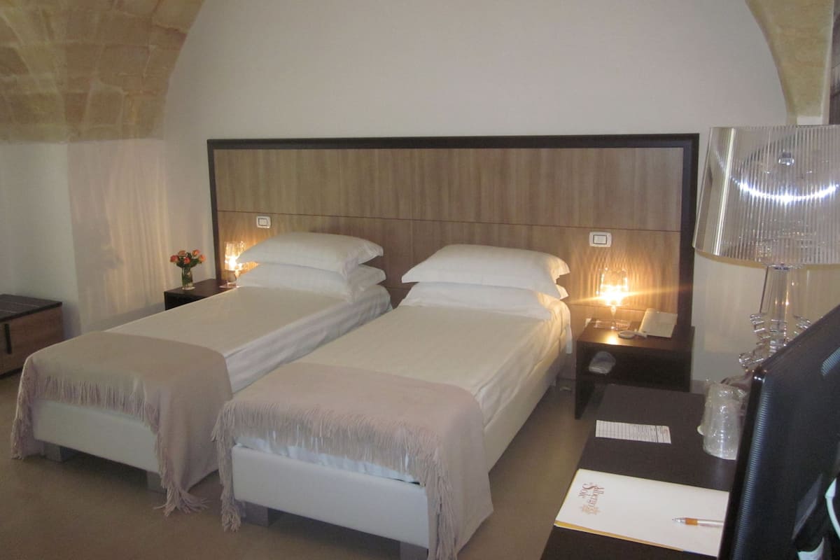 Habitación de hotel con dos camas, una mesita de noche con lámpara, un escritorio con computadora y una pared de piedra.