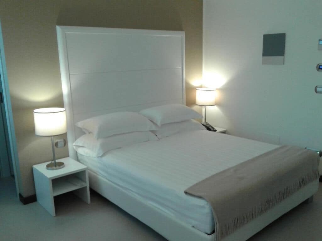 Een netjes opgemaakt bed met witte lakens en kussens, een nachtkastje met een lamp, en een wandlamp.