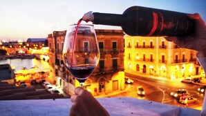 Una persona sirve vino de una botella en una copa, con un paisaje urbano y edificios al fondo.