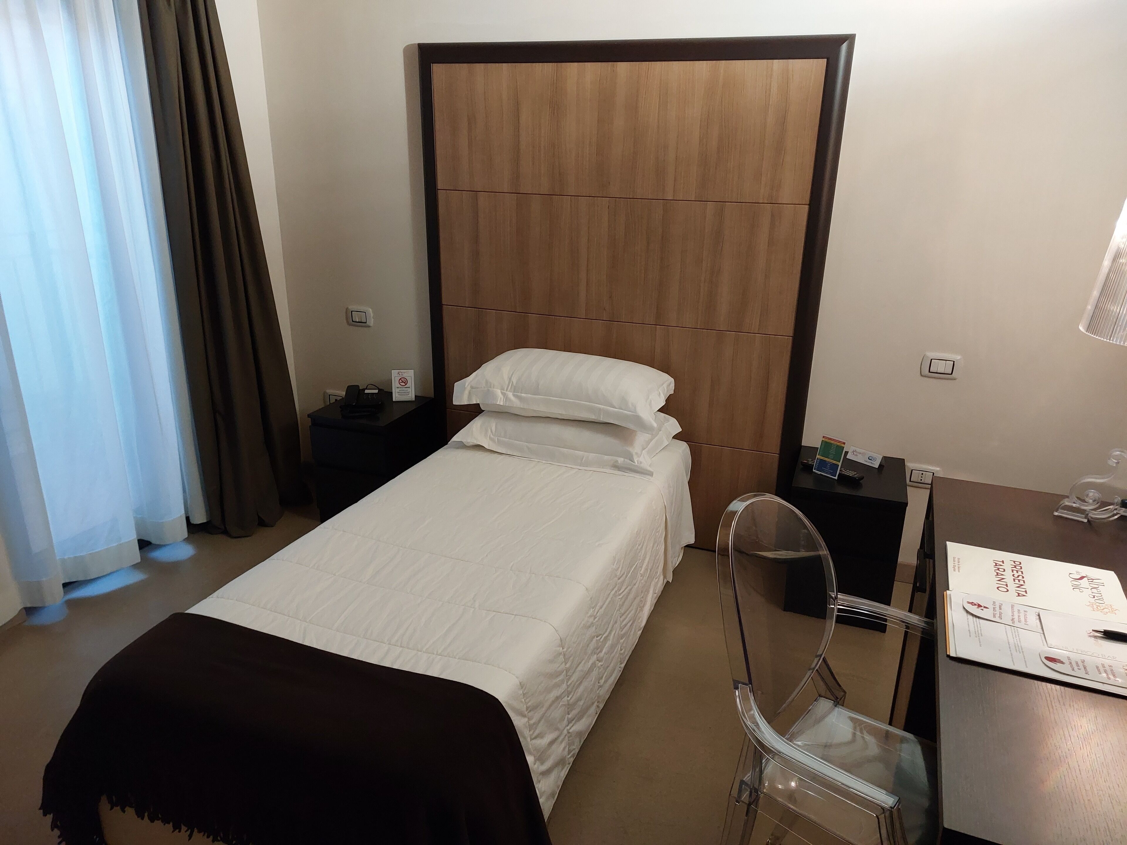 A hotel room with a bed, a desk with a chair, a nightstand, and a wardrobe.