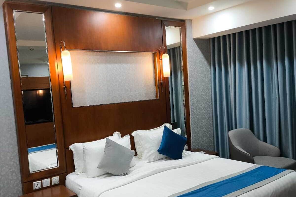 Quarto de hotel com uma cama grande, mesa de cabeceira, cadeira, espelho e janela com cortinas.
