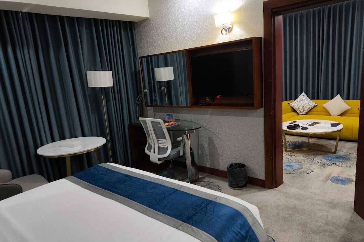 Quarto de hotel com cama, escrivaninha, cadeira, mesa pequena, televisão e sofá amarelo com almofada estampada.