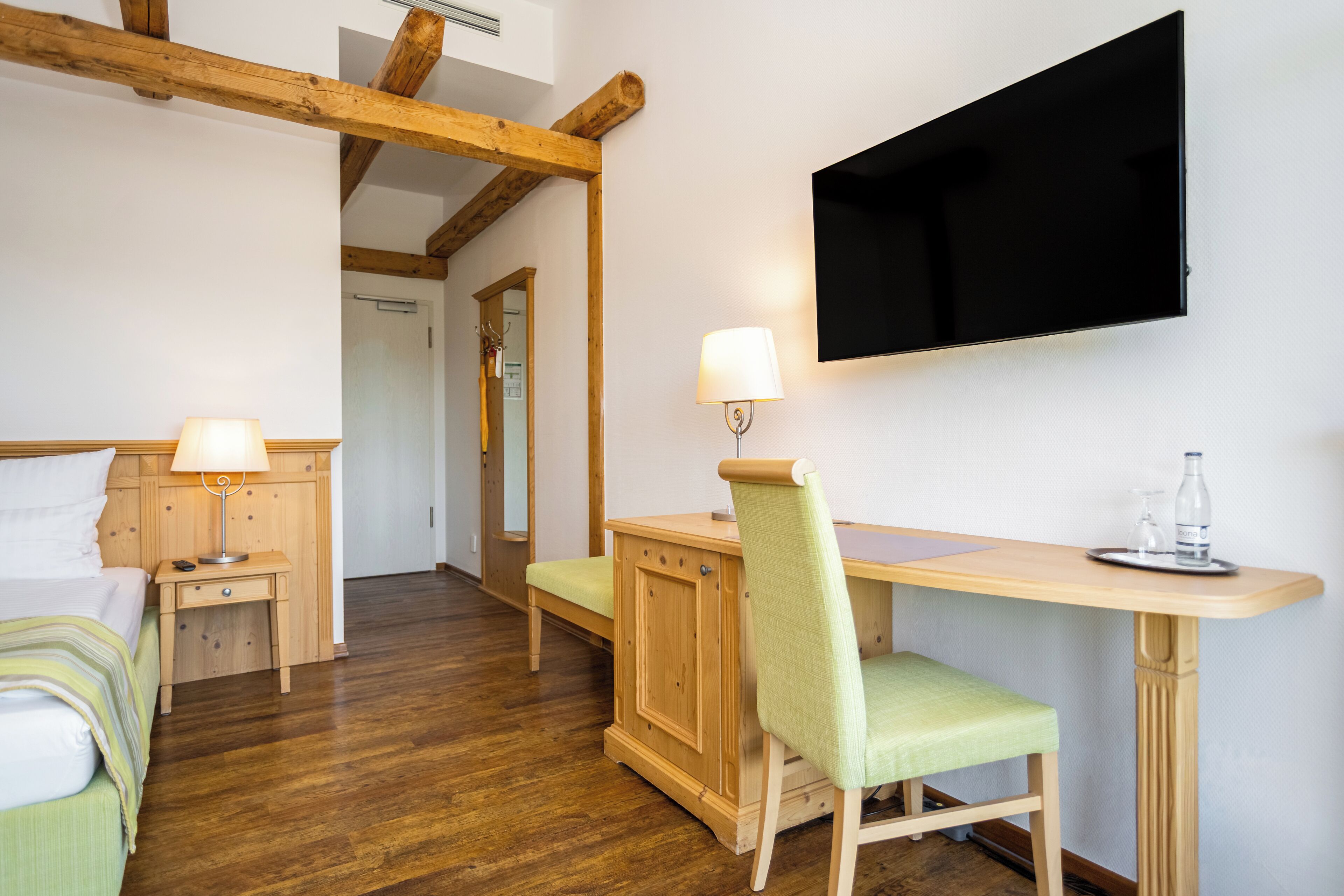 A room with a bed, a desk, a chair, a TV mounted on the wall, and a wooden floor.