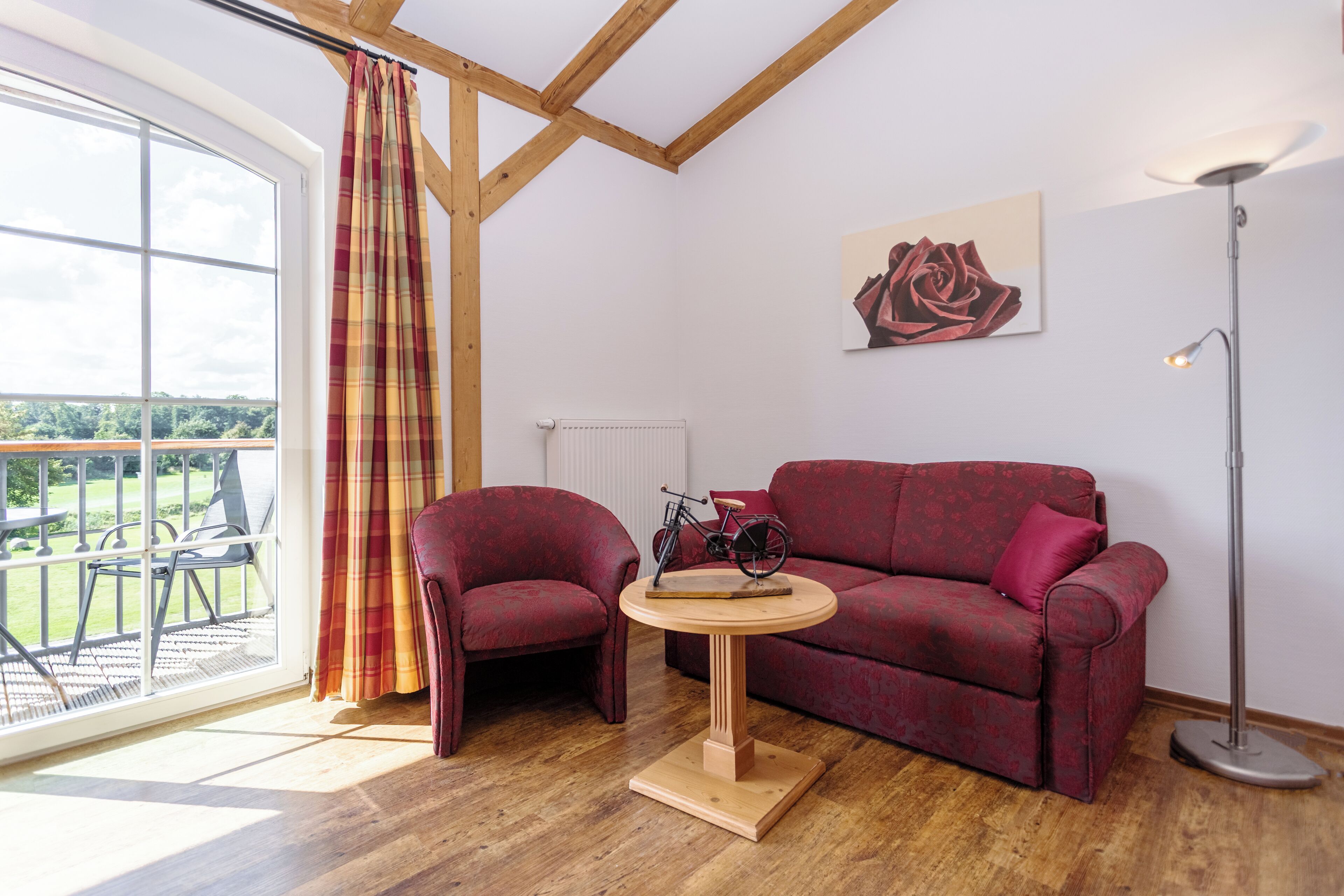 A room with a red sofa, two armchairs, a round wooden table, a bicycle, a floor lamp, a large window with curtains, and a painting of a rose.