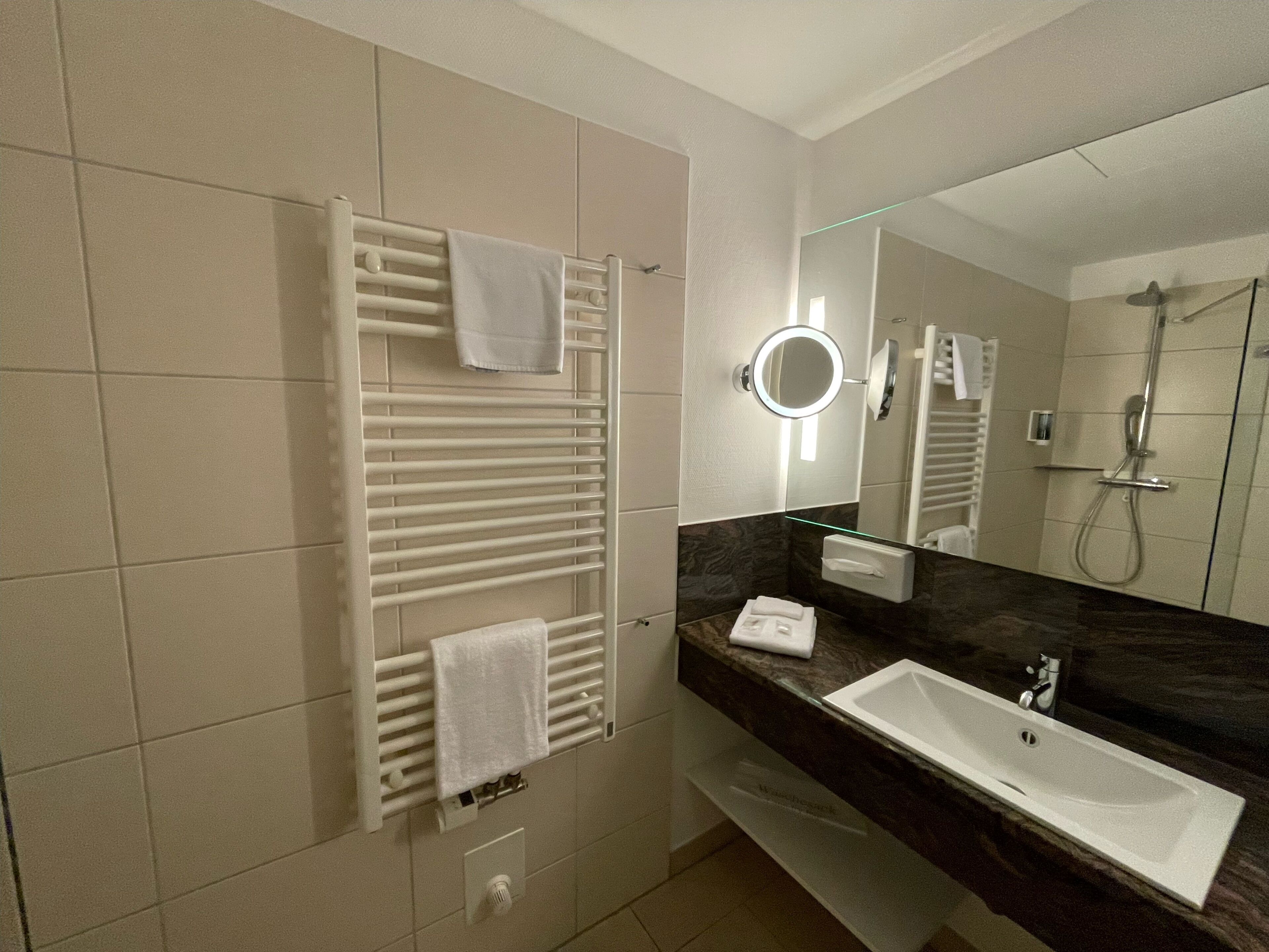 Ein modernes Badezimmer mit integriertem Handtuchhalter, einem runden Spiegel mit Beleuchtung, einem Waschbecken und einer Duschkabine.